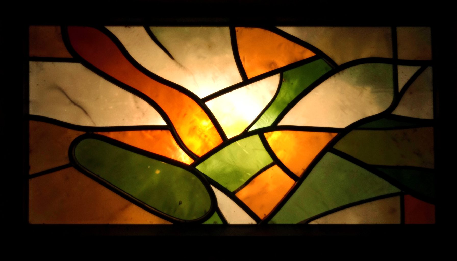 A stained-glass composition with bold leaded segments in warm amber, olive green, and cream tones, forming an abstract organic pattern with a soft glowing light behind it